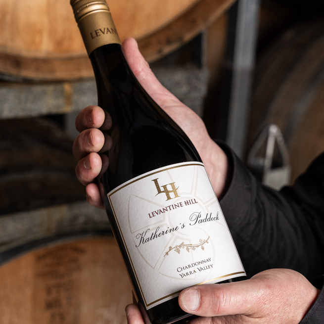 Hand holding a bottle of Levantine Hill Katherine's Paddock Chardonnay wine with barrels in the background