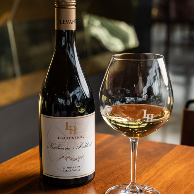 Bottle of Levantine Hill Katherine's Paddock Chardonnay with a glass of wine on a wooden table.